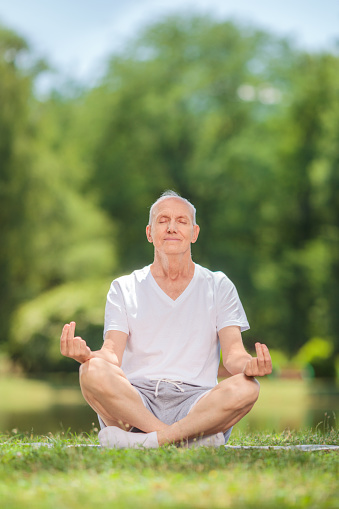 Vertical shot of a senior man meditating seated on blanket in a field by a pond