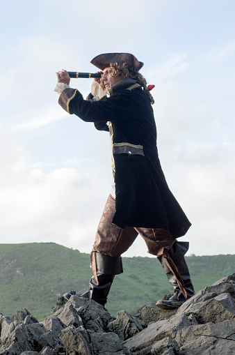 Vertical image of a mature man dressed as a pirate in a traditional costume, standing on  a rocky area on the beach, with cliffs in the background. Looking through a telescope.
