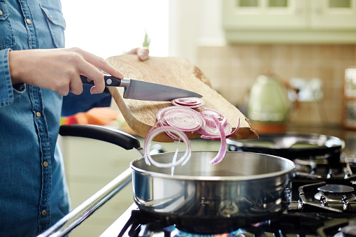 Midsection image of woman pouring chopped onions in saucepan. Utensil is placed on gas stove. Close-up of female holding cutting board and kitchen knife. She is preparing food in domestic kitchen.