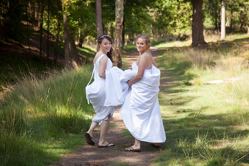two brides walk on forest path with skirts in their arms while looking back smilingin their arms