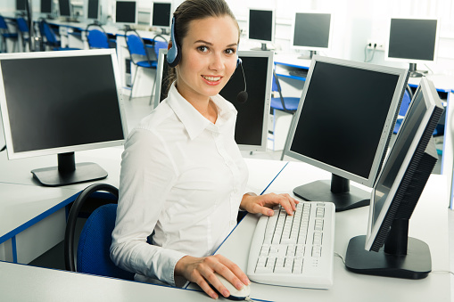 Image of pretty customer service representative wearing headset sitting at computer and looking at camera with smile   