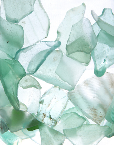 Pieces of aqua sea glass are illuminated from behind by light.  There are varying tones, edges and textures to the sea glass. Pieces of turquoise sea glass have a strong highlighted background in a macro view.  The layers produce different tones and hues of color.  The perspective is such that the sea glass looks to be coming toward the viewer.  Sea glass pieces of different hues of aqua look as though they are floating in the air.  Approximately 15 pieces are in a tight cluster, overlapping, with a highlighted background. The pieces have different textures and random shapes including ridges, and lines. As if floating, stacked blue sea glass of varying shades are against natural back light. Aqua sea glass, azure sea glass, turquoise sea glass are in various stages of wear.  The sea glass pieces are different sizes and perspective, some facing full front while others are a on their sides.  The natural effects of the ocean on old pieces of colored glass is photographed with a Canon macro lens showing a central focus.
