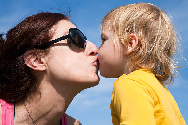 two years old age blonde baby yellow shirt kissing in mouth to brunette woman mother sunglasses with blue sky background