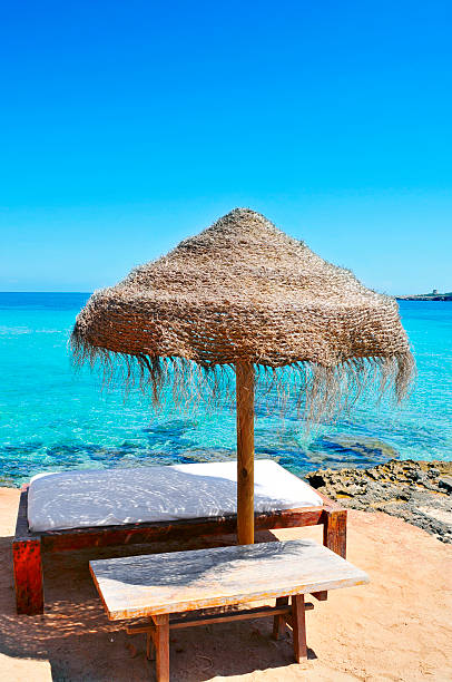 detail of a relaxing area in a beach in Ibiza, Spain, with a comfortable sunlounger and a rustic umbrella made of natural fibers