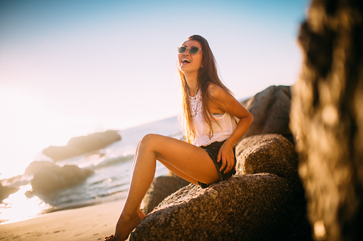 Pretty teen Asian girl sitting on some rocks laughing out loud while enjoying a summer afternoon at the beach