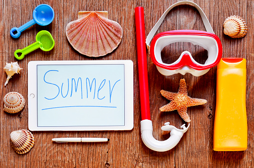 high-angle shot of a rustic wooden table full of summer stuff, such as a starfish, some seashells, a diving mask and a snorkel, a bottle of sunblock and a tablet with the word summer written in it