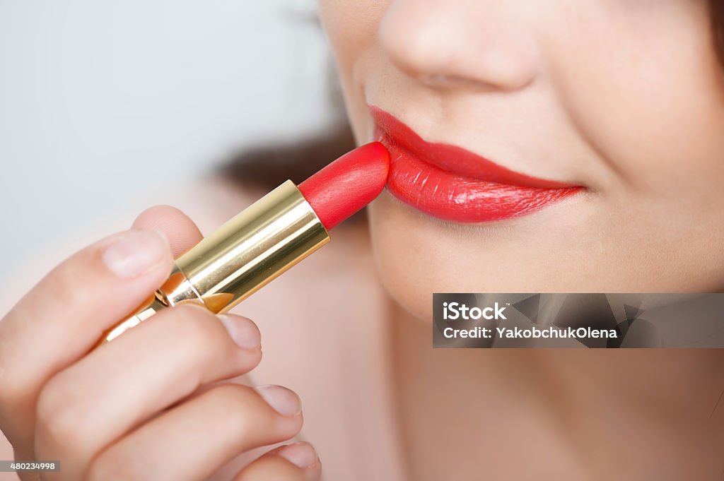 Cheerful young girl is doing her make-up Close up portrait of attractive lips of beautiful woman. She is rouging her lips with red lipstick. The lady is gently smiling. Isolated on grey background Lipstick Stock Photo