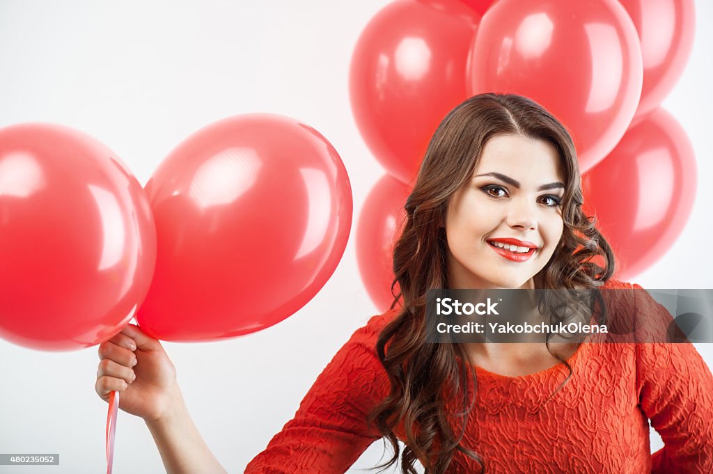 Pretty young woman in red dress is giving present Cheerful girl is holding red balloons and resenting it to camera. She hides another balloons behind her back. The girl is smiling secretly. Isolated on grey background 2015 Stock Photo