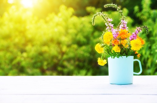 bouquet of wild flowers in a cup on a wooden table on a background of greenery and sunlight