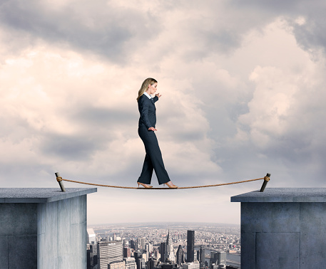 A businesswoman balancing on a tightrope high above a big city. She looks down at the rope before her next step as she nears the midpoint of the tightrope.  Metal spikes are anchored into the concrete to help support the tightrope.  The New York City skyline can be seen in the distance as a fog persists over the buildings.