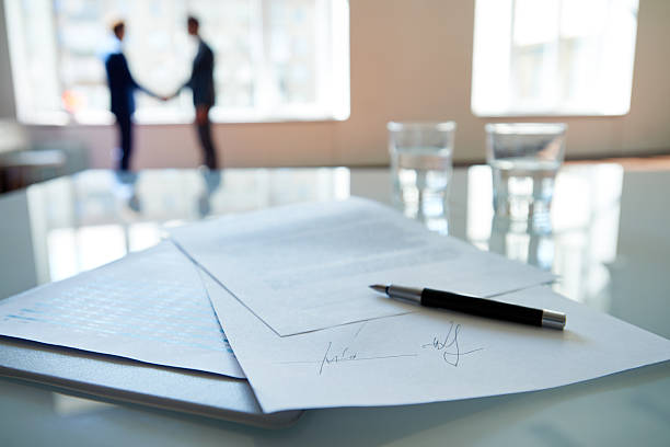 Business contract Signed business contract lying on table, business partners shaking hands in the background agreement stock pictures, royalty-free photos & images