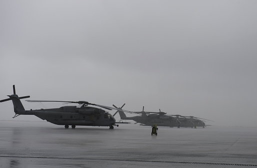 A line of four Marine MH-53 Helicopters sit on a flight line in a grey rainstorm