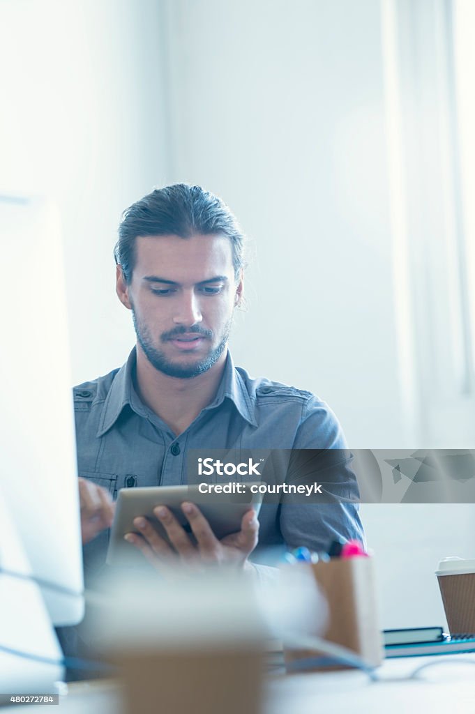 Modern businessman using digital tablet Modern businessman using digital tablet with coffee. He is sitting at his desk with a computer, and digital tablet. He is sitting at a large table which has other technology on it. Could be a graphic design studio or small business. He could be surfing the web or looking at social media or email. There is a large window behind him. Copy space 20-29 Years Stock Photo