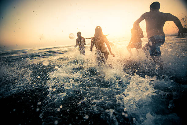 Group of friends running into the water Group of friends running in the ocean at sunset and having fun people jumping sea beach stock pictures, royalty-free photos & images