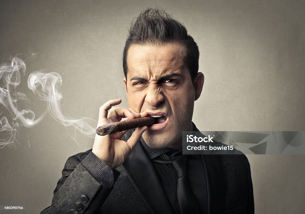 Cigar A man, wearing jacket, tie and shirt total black, is smocking a big cigar and is knitting his nose and his forehead as the smoke was too much. He's looking at the camera. The background is grey. Men Stock Photo