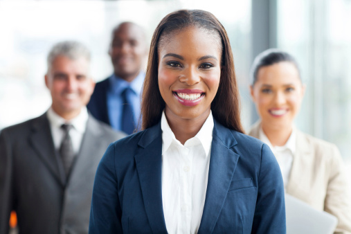 Portrait of confident businesswoman sitting in the office. Smiling female team leader, looking at camera. Behind of her colleagues in board room. Multi-ethnic group of business people on meeting in conference room.