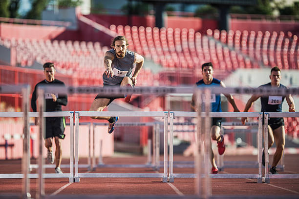 Group of male athletes jumping hurdles on a race. Four young man jumping hurdles on a sports race at stadium. hurdling track event stock pictures, royalty-free photos & images