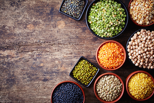 Bowls of various legumes (chickpeas, green peas, red lentils, canadian lentils, indian lentils, black lentils, green lentils; yellow peas, green mung beans) on wooden background