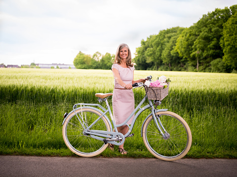 Full length portrait of a beautiful mature woman standing alongside her vintage bicycle on a country road in a lush green park