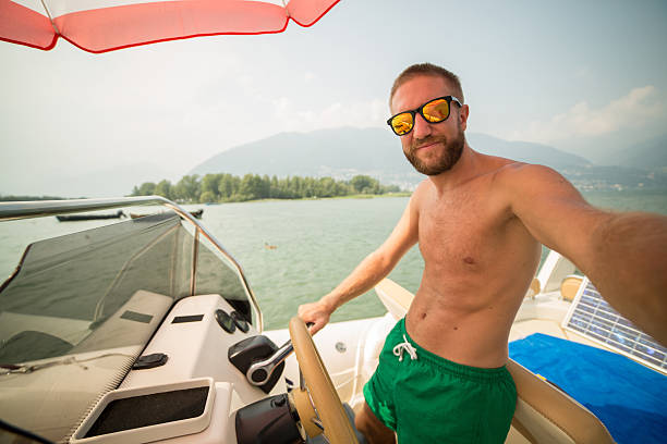 Young man on a boat taking a selfie while driving it, he is using a smart phone. Sunny summer day.