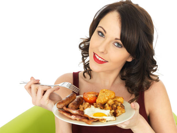 Young Attractive Woman Eating a Full English Breakfast, looking at the camera smiling and happy with her head leaning slightly to one side, about to eat and enjoy the cooked fried food, isolated against a plain white background