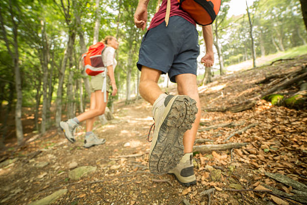 Hikers walk on a mountain trail. The focus of the image is on one hiker's left boot. The sole of the boot is in the foreground of the image. The hiker wears sports clothing and a backpack. Another hiker is visible in the background, walking on the dirt trail.