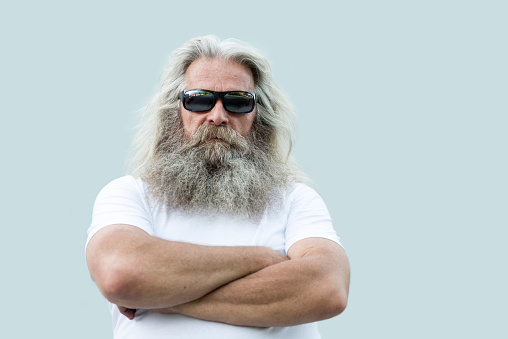 A portrait of a senior man with long grey hair and a long beard standing in front of a  wall with his arms crossed. He is wearing a white shirt, dark sunglasses  and he is looking straight and serious at the camera. There is some copy space on the right side of the photo.