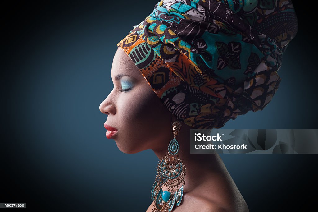 young beautiful fashion model with traditional african style young beautiful fashion model with traditional african style with scarf, earrings and makeup on dark blue background.   African Ethnicity Stock Photo