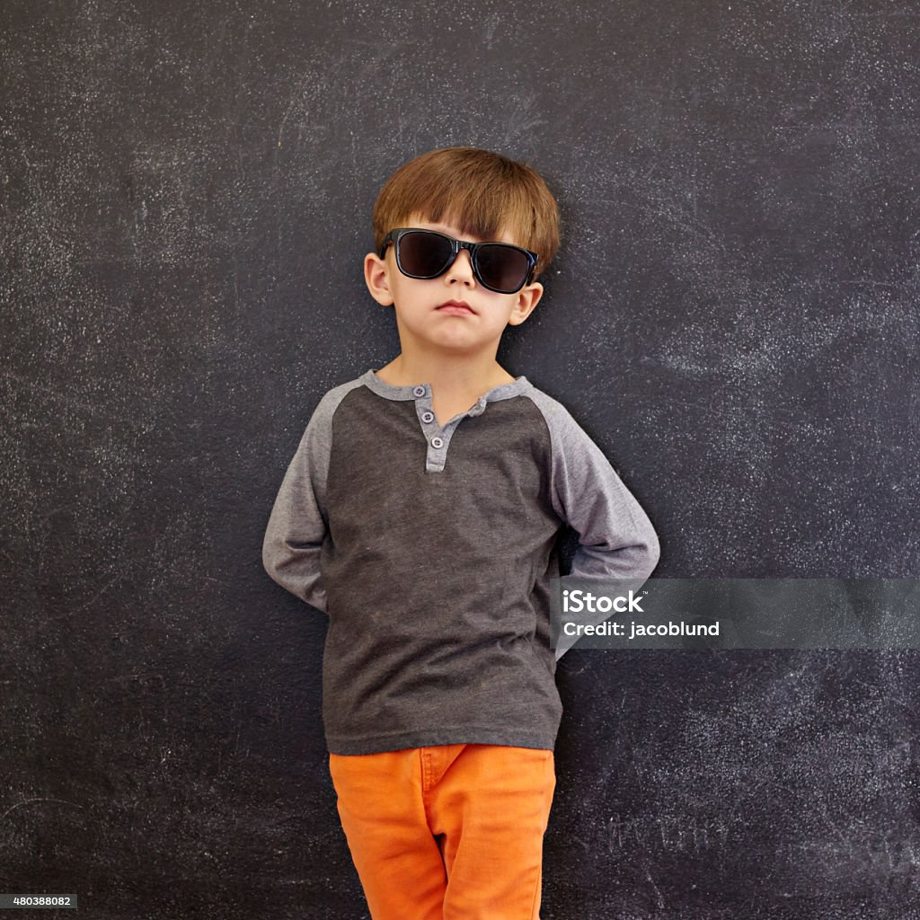 Stylish little boy wearing sunglasses leaning on a blackboard Cute little kid standing in front of the blackboard. Stylish little boy wearing sunglasses leaning on a blackboard looking at camera. 2-3 Years Stock Photo