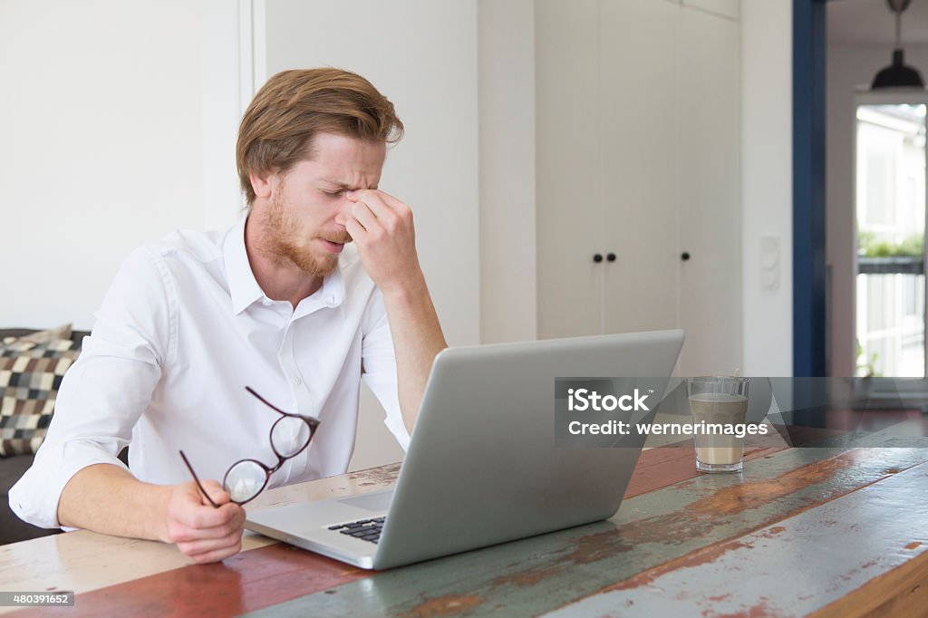 young man sitting with laptop and a headache young red-haired man sitting at table with his laptop and having a headache 2015 Stock Photo