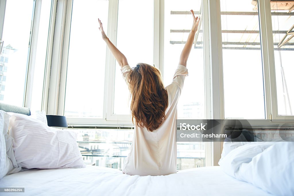 Morning stretch in bed Young woman is doing morning stretching in bed, arms raised, rear view Waking up Stock Photo