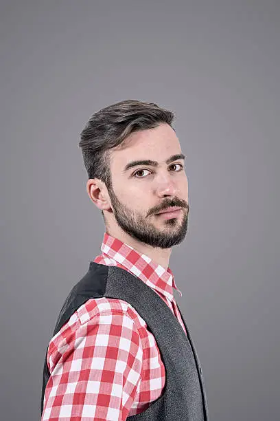 Desaturated studio portrait of relaxed young bearded hipster side view looking at camera over dark grey background with vignette.