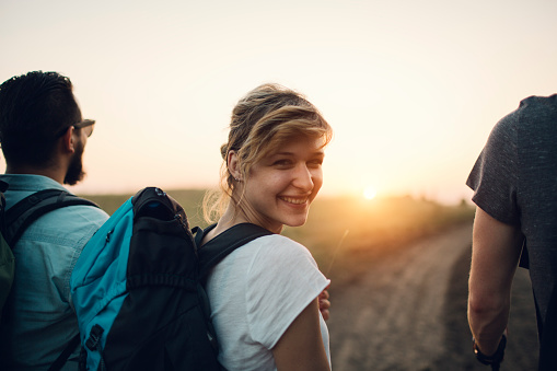 Group of young people hiking together. Closeup of an young smiling woman looking at camera over shoulder. They are walking to sunset.