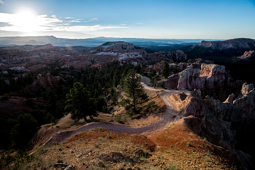 The sun peaks over the distant mountains illuminating the Bryce Canyon National Park. A deserted path leading into the valley guides tourists into an alien world of hoodoos, cliffs, and grand vistas, all formed by relentless nature, erosion, and time.