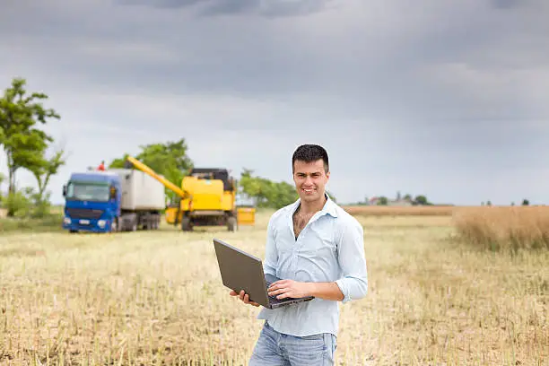 Young attractive farmer with laptop standing in rapeseed field, truck and combine harvester working in the field in background