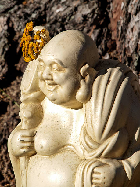 Laughing garden Buddha statue with flowers Porcelain laughing Buddha garden statue with marigold flowers over his ear, setting under a pine tree in the sunlight. laughing buddha garden statue stock pictures, royalty-free photos & images