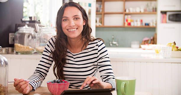 Young Woman Eating Breakfast Whilst Using Digital Tablet Young Woman Eating Breakfast Whilst Using Digital Tablet medium shot stock pictures, royalty-free photos & images