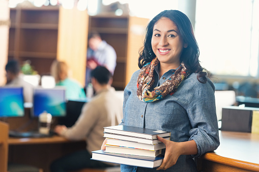 Young adult Hispanic woman is college student continuing her education. She is looking at the camera and smiling while she holds a large stack of college textbooks in library. She is borrowing books from school library. Woman is standing near checkout counter in front of computers and large bookshelves. She is wearing casual clothing with a colorful scarf.