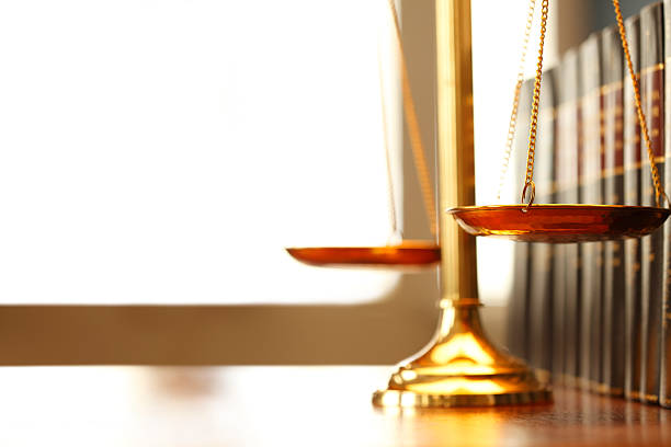 A justice scale on a desk next to a row of law books. Light filters in through a window in the background creating ample negative space for coy.  Photographed using a very shallow depth of field.