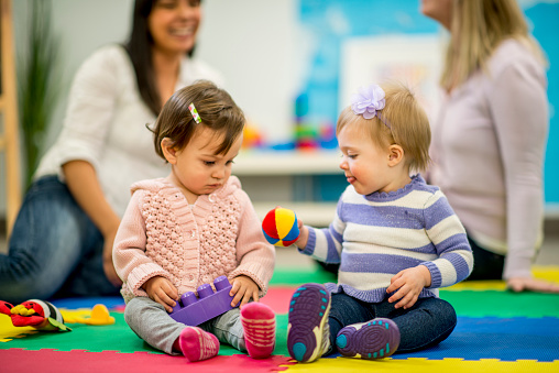 A multi-ethnic group of babies, their mothers in the background, playing together with toys in a daycare / preschool setting.