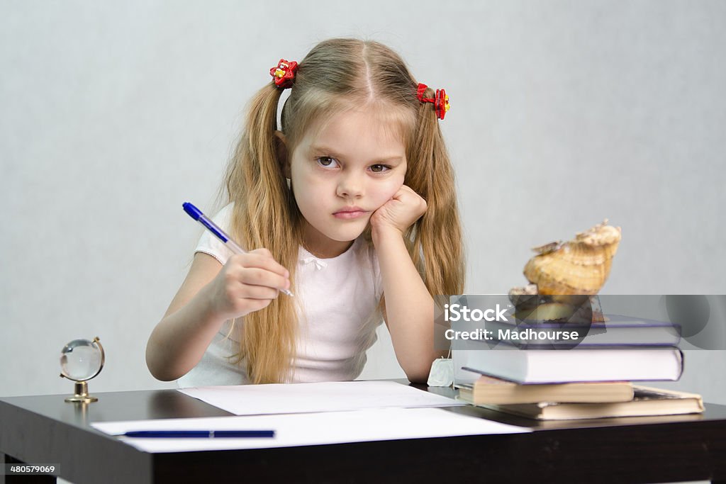 child at the lessons at the table girl sits at a table and wrote on a sheet of paper. There are tombs of books, sheets of paper, a pen, glass globe, with a glass of tea, sea shell. The image of the writer Book Stock Photo