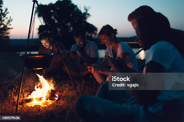 Hikers Using Smart Phones Around Campfire Stock Photo - Download Image Now - Camping, Campfire, Cooking