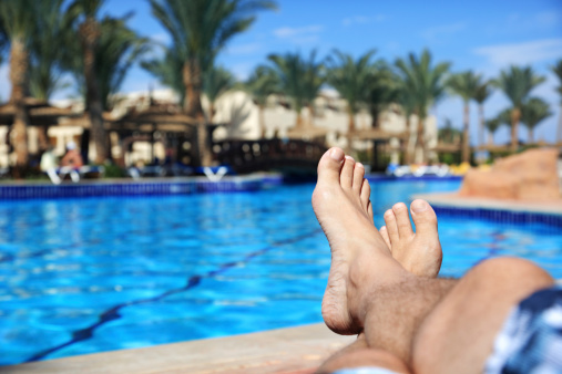 Sunbathing by the hotel tourist resort swimming pool, mans legs lying down on a sunlounger looking over the water