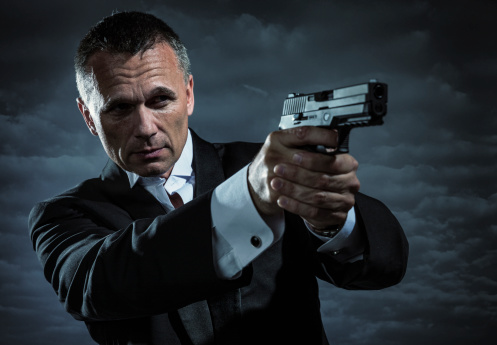 A handsome mature male secret agent, bodyguard, spy, or security staff dressed in an elegant tuxedo and bow tie points a modern pistol on a stormy night with cloudy sky in the background.