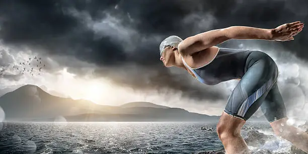 Female swimmer / triathlete standing in the sea about to dive in and swim. The athlete is set in a generic seascape, by mountains under dramatic stormy evening sky at sunset. Swimmer is wearing generic unbranded swimsuit, swimming cap and goggles. With intentional light flare and bokeh water spray effects. 