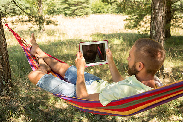Man relaxing on hammock in summer using digital tablet Young man relaxing on hammock in summer using a digital tablet. hammock men lying down digital tablet stock pictures, royalty-free photos & images