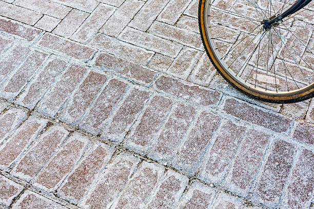 An old bicycle with a corroded metal wheel rim, rusty spokes and a brittle, cracked rubber tire is parked on a heavily salted and winter stained urban sidewalk.
