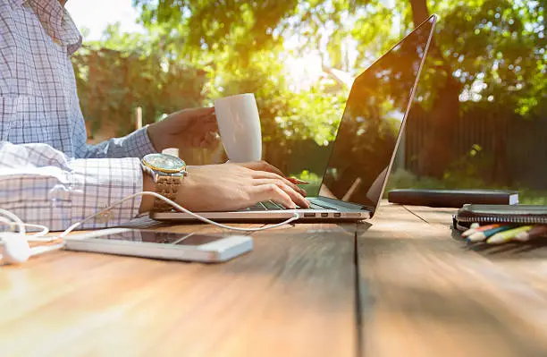 Business style dressed man sitting at natural country style wooden desk with electronic gadgets around working on laptop drinking coffee sunlight and green terrace on background