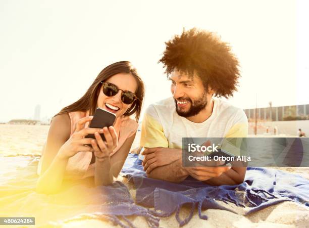 Young Couple Lying On Blanket On Beach Using Smart Phone Stock Photo - Download Image Now