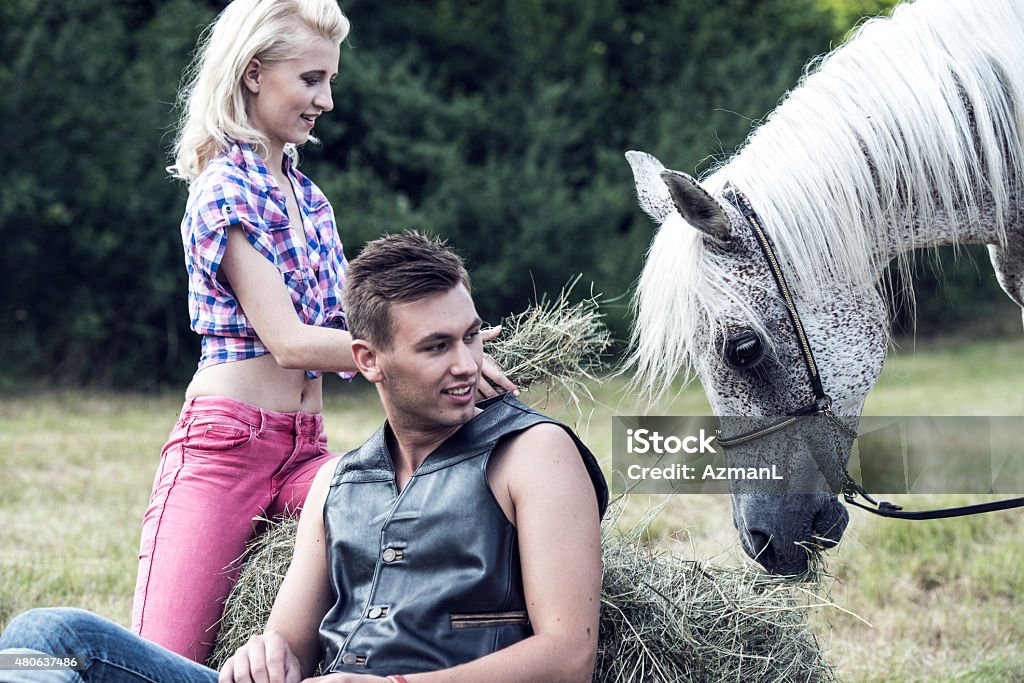 Cheerful Couple and a Horse Cheerful young couple sitting on a meadow, enjoying nature, relaxing. Woman is feeding a horse. They are both happy. Adolescence Stock Photo